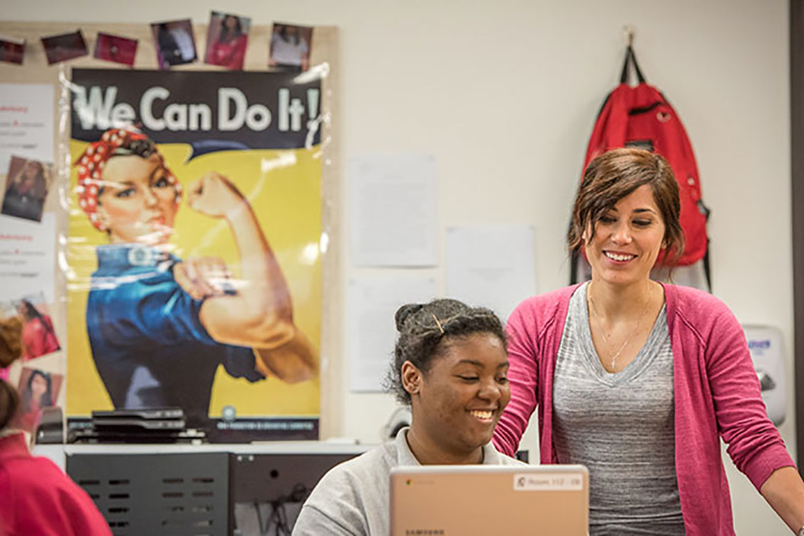 A young woman demonstrates how to become a high school teacher by helping a student complete schoolwork on a computer in the classroom.