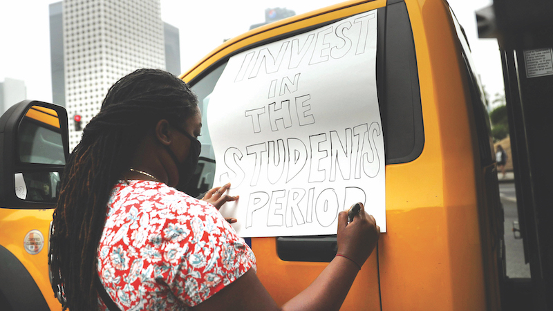 Image of a woman at a protest writing the words &quot;Invest in the students, period&quot; on a poster board sign.
