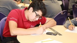 A high school student writes at his desk in a classroom, and is engaged in curriculum that follows K-12 content standards