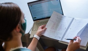 A student engages in a virtual classroom her desk, watching a laptop screen while working in a notebook
