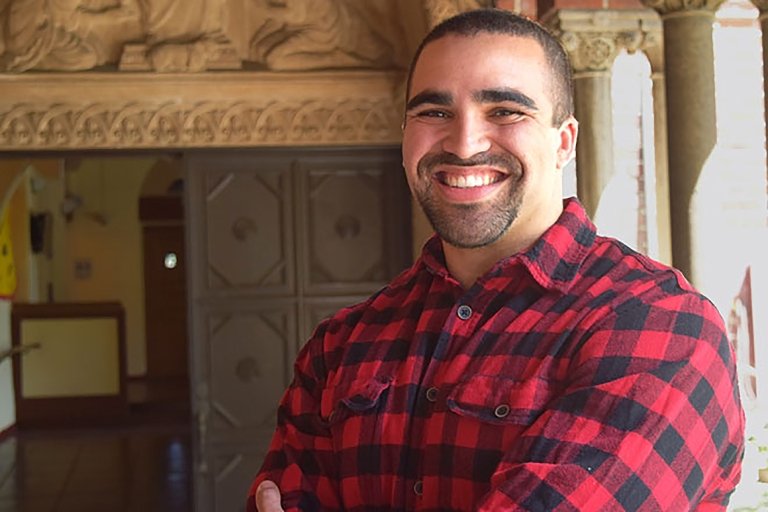 Steven Spencer, MAT &#039;18 is a high school teacher who poses for a photo in a checkered shirt on a college campus
