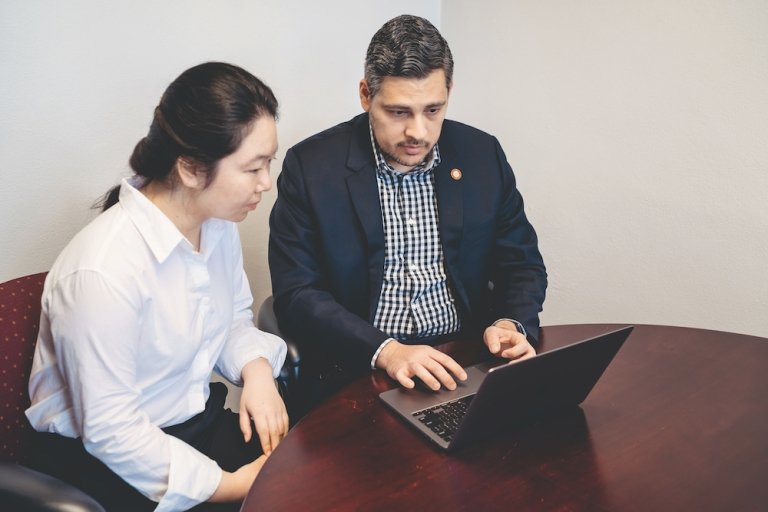 Changzhao Wang and Stephen Aguilar look at a laptop computer together. They are seated a wooden table. Changzhao Wang is wearing a white shirt and Stephen Aguilar is wearing a blue blazer over a plaid shirt.