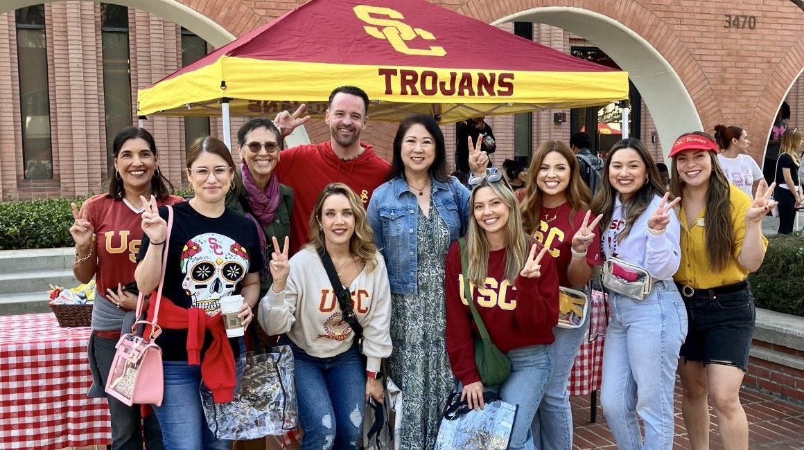 Group of USC alumni and staff posing with peace signs under a USC Trojans tent
