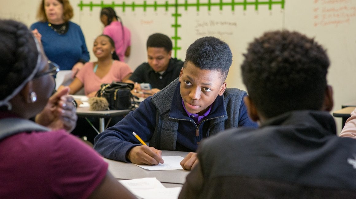 Student writing at a desk while participating in a classroom group discussion