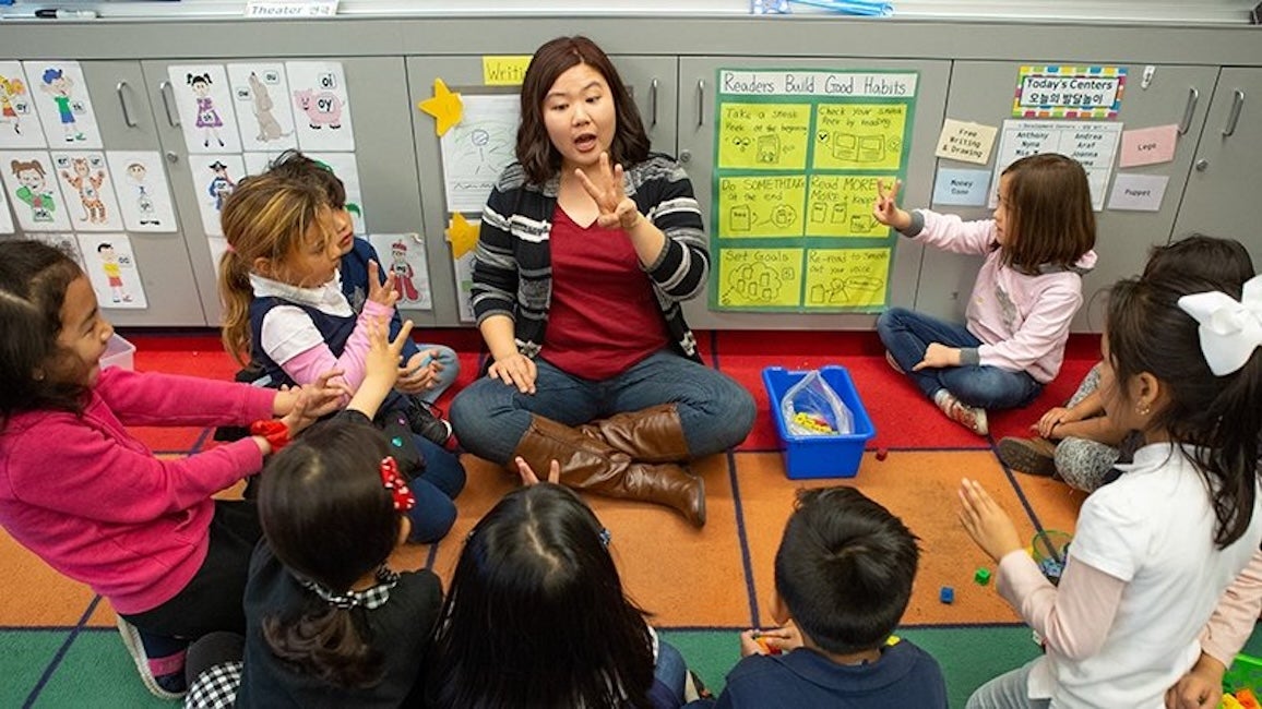 Teacher sitting on the floor making hand gestures while surrounded by young students
