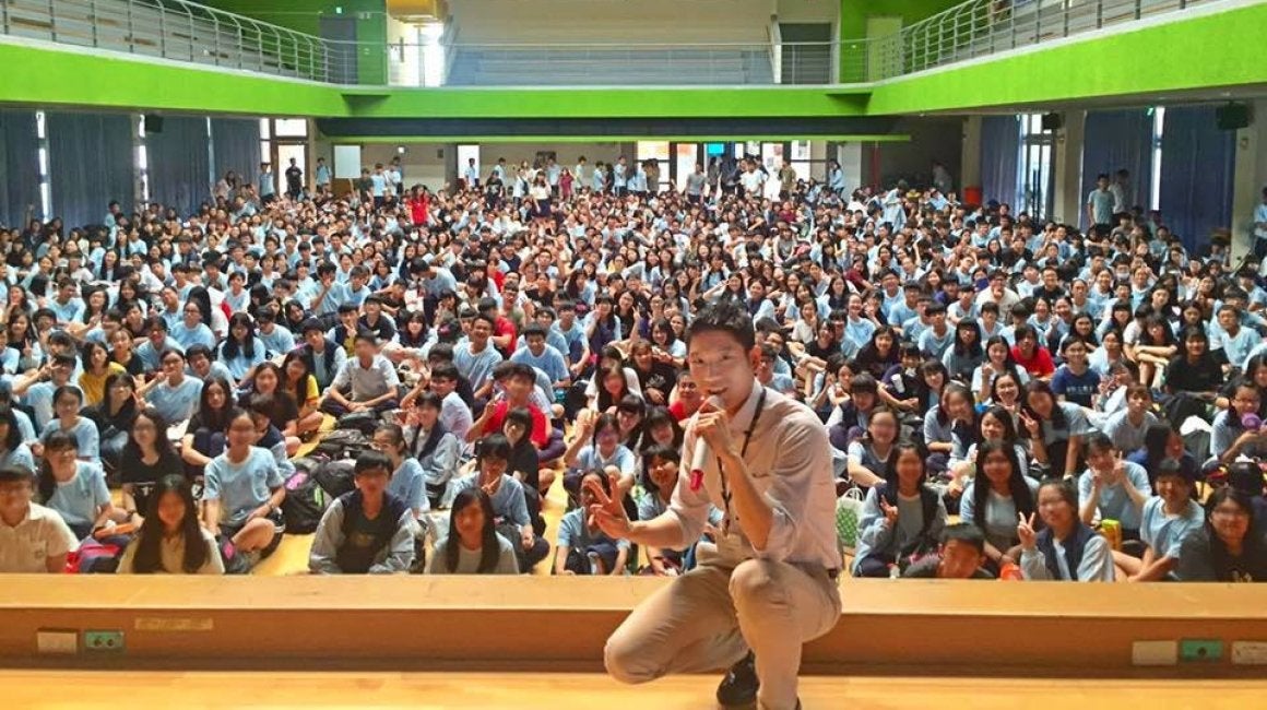 Eric Yang crouching and speaking into a microphone in front of a large auditorium of students