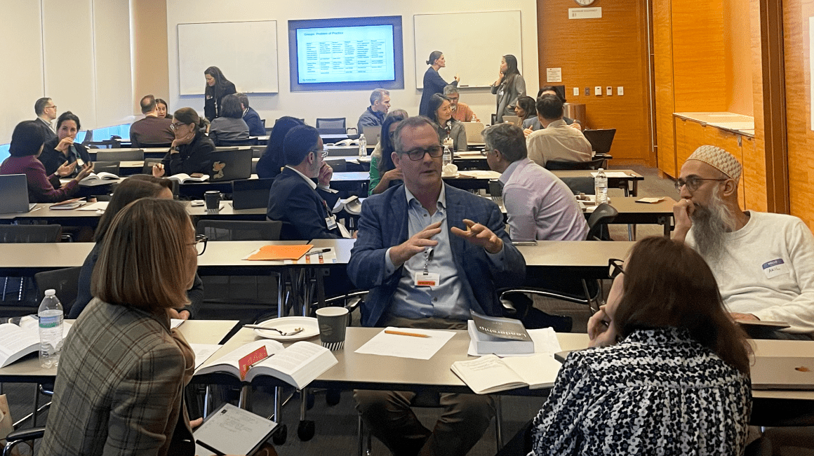 Man gesturing while talking at a conference table surrounded by attendees