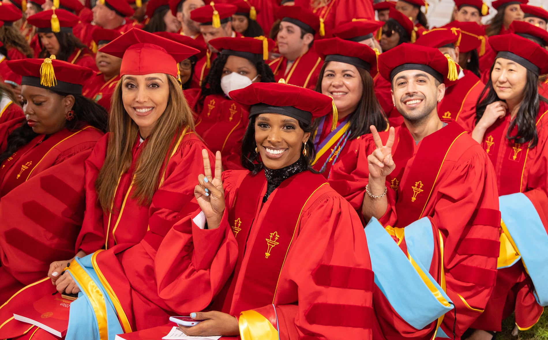 Group of doctoral graduates in red and gold regalia smiling and giving peace signs