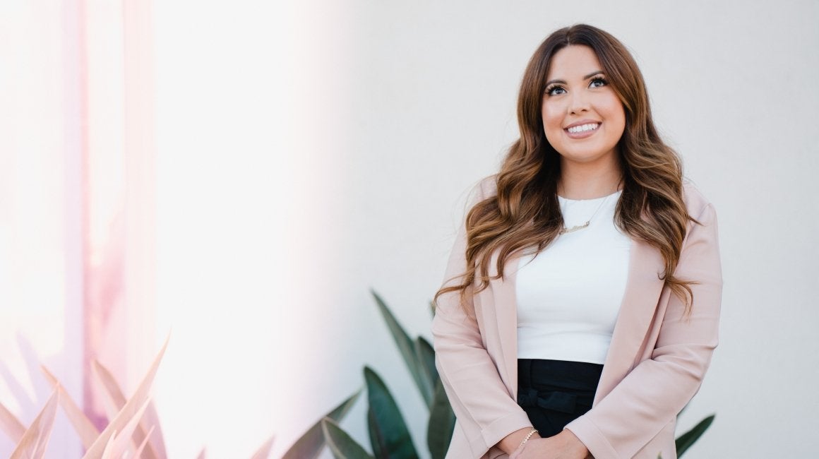 Headshot portrait of a woman smiling in a pink blazer outdoors near plants