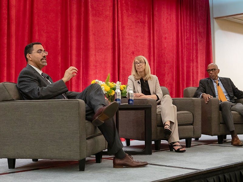 Three panelists seated in armchairs on a stage during a discussion in front of a red curtain