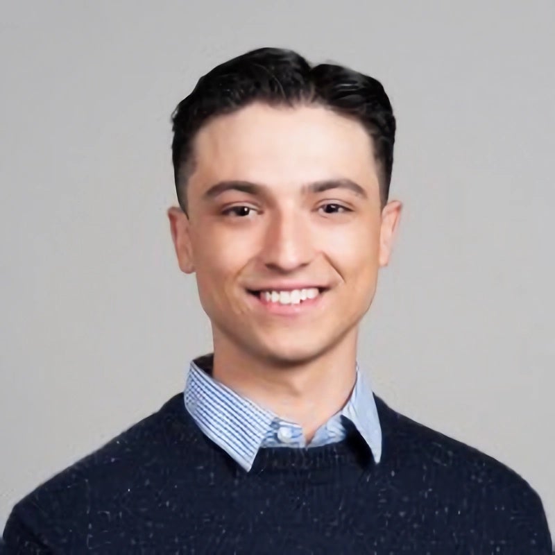 Headshot portrait of Moises smiling in a dark sweater against a gray background