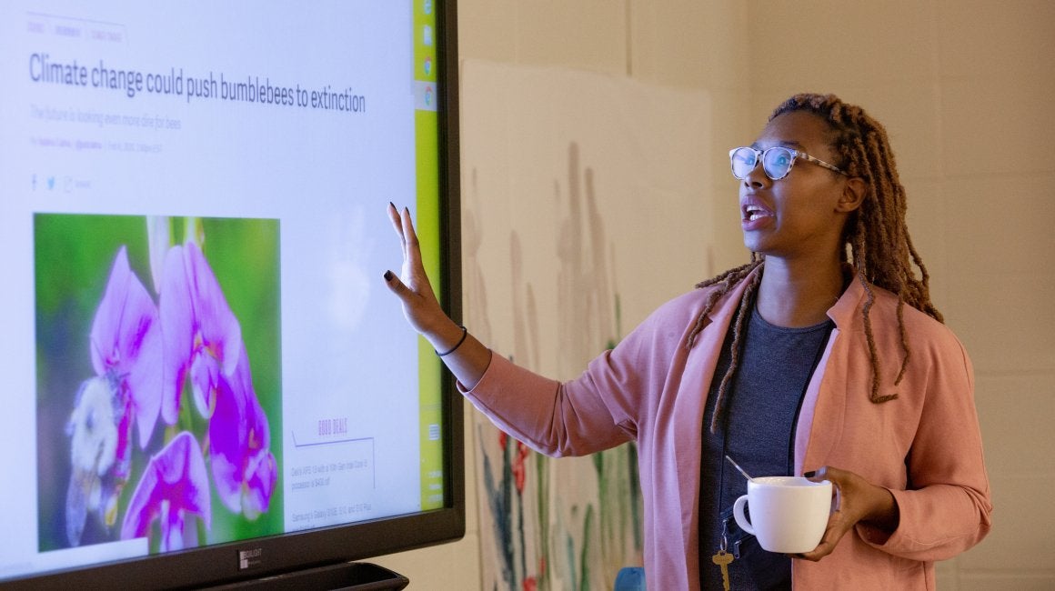 Teacher gesturing while presenting a climate change news article on a classroom smartboard, holding a coffee mug