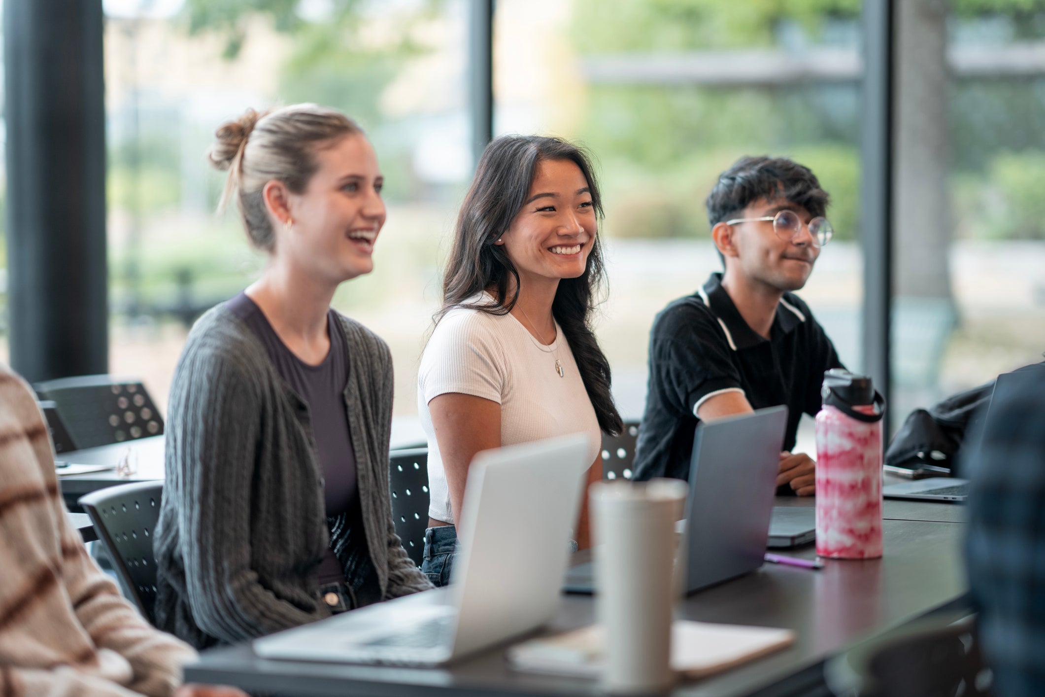 Three students smiling and laughing together at a table with laptops near large windows