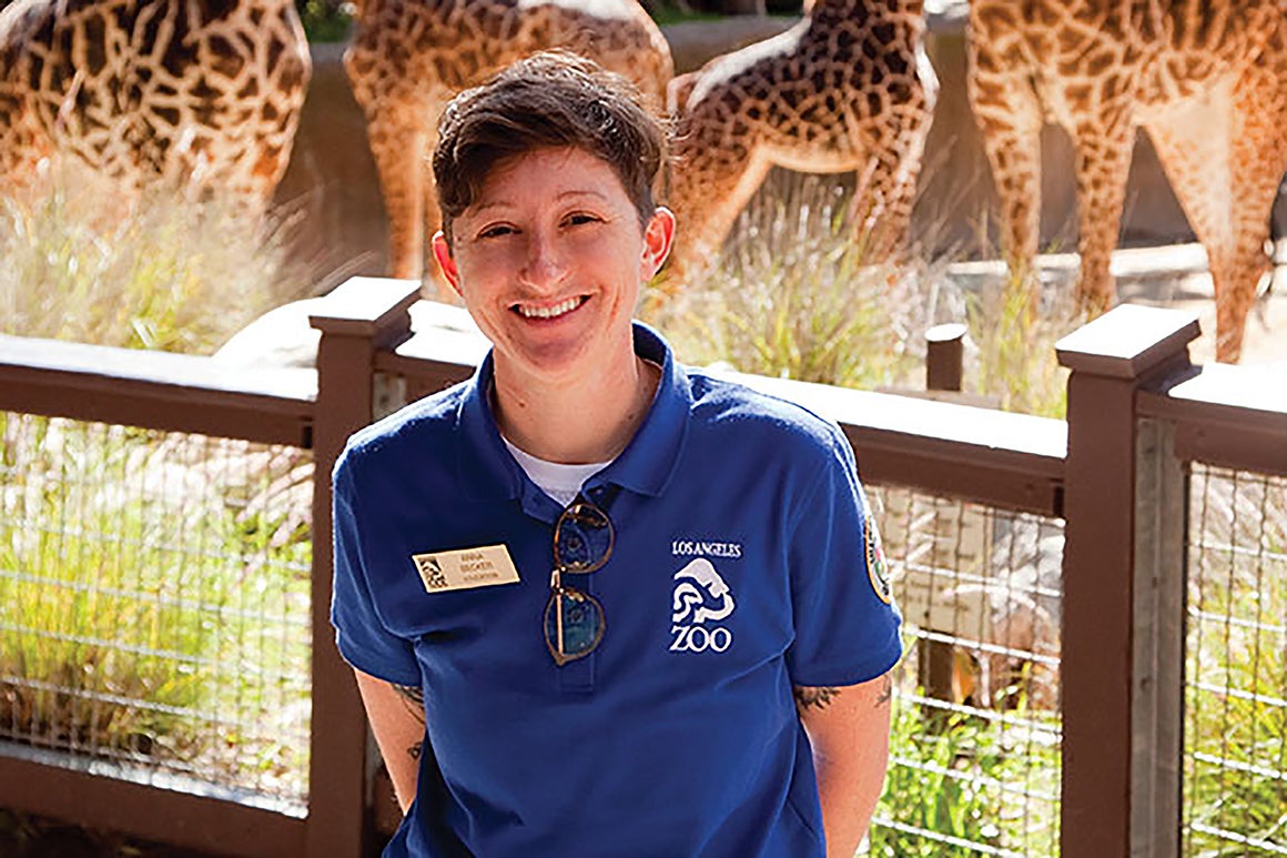Anna Becker, MAT '18, a teacher, smiles in front of giraffes at her workplace: The Los Angeles Zoo